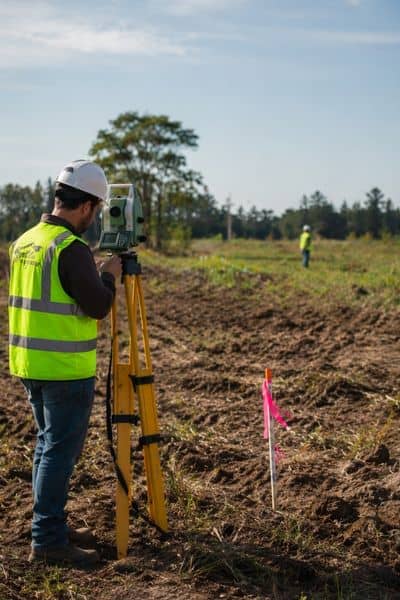 Surveyor in a high-visibility vest and hard hat uses a tripod-mounted instrument in a dirt field; pink flag marker and another worker in the background.