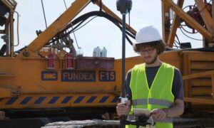 Land surveyor using GNSS equipment beside construction machinery, reflecting fieldwork associated with an as-built survey
